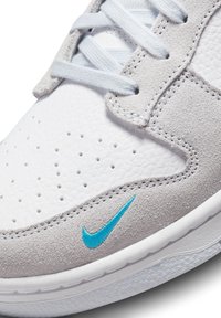 Vita lädersneakers med grå mockadetaljer, blå broderad Nike-logotyp, perforerat tårum och ljusblå snörningar.