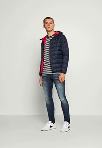 Gewatteerde marineblauwe jas met een rode voering, gedragen over een gestreepte t-shirt, gecombineerd met slim-fit blauwe jeans en witte sneakers.
