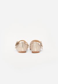 Beige achteraanzicht van platte schoenen met een geweven textuur, metalen accenten en een bloemenstrikdetail. Rubberen zool en verstelbare band.