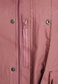 Gros plan sur une veste mauve montrant une fermeture éclair centrale, une plus petite fermeture éclair verticale de poche, et des boutons-pression en métal sur le tissu.