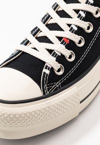 Czarne trampki na płaskiej podeszwie z gumowym noskiem, beżowymi sznurówkami, srebrnymi oczkami i fakturowaną białą podeszwą z wzorem w romby.