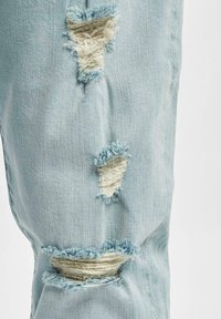 Tissu en denim bleu clair avec trois déchirures verticales usées laissant apparaître des fils blancs effilochés en dessous.