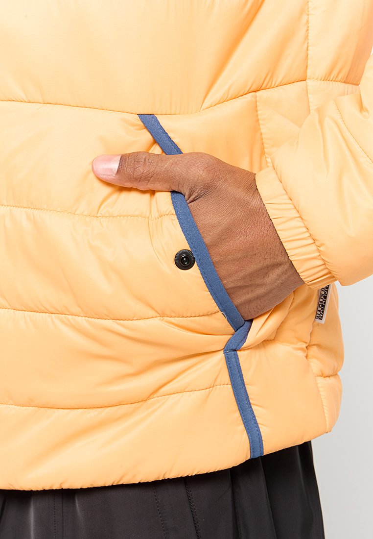 Giacca a vento in morbido tessuto giallo con sezioni cucite, piping a contrasto blu e una tasca frontale con chiusura a bottone a pressione.