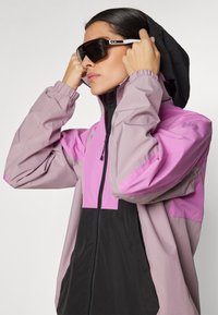 Chaqueta ligera en púrpura y negro, con cremallera completa, capucha ajustable y puños elásticos. Llevada con gafas de sol negras.