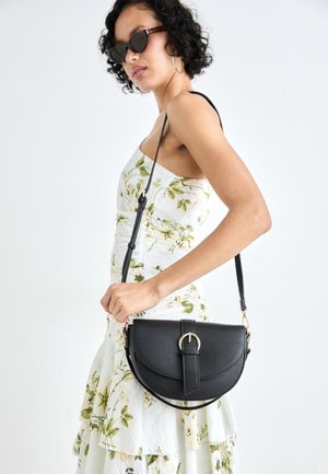 Kvinde iført solbriller og en blomsterkjole, der holder en sort læder crossbody taske med en guldbuckle mod en hvid baggrund.