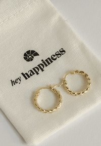 Pendientes de aro de oro con un diseño retorcido, exhibidos sobre una funda beige que lleva el texto "hola felicidad". Apariencia simple y elegante.