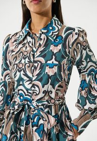 Blumenmuster-Shirtkleid mit Knopfleiste, klassischem Kragen, Rüschensäumen und einem gebundenen Taillengürtel. Die Farben sind Teal, Pink und Cream.
