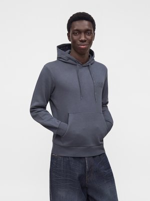 Ung mand iført grå hættetrøje med forlomme og blå jeans, står med hænderne i hættetrøjens lomme, neutral hvid baggrund.