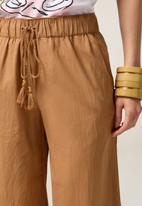 Pantalons larges, légers et bruns avec une taille élastique et un cordon de serrage. Comprend des pompons avec des accents dorés. Texture lisse.