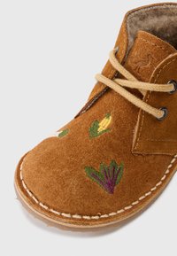 Brązowe zamszowe buty z beżowym sznurkiem, haftowane warzywa, widoczne szycie i miękka podszewka.