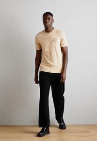 Helles beige T-Shirt mit kurzen Ärmeln und einem kleinen roten Grafikdesign auf der linken Brust, kombiniert mit schwarzen Hosen und schwarzen Lederschuhen.
