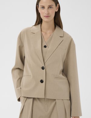 Kvinde iført beige skræddersyet blazer og matchende plisserede bukser, stående mod en ensfarvet lys baggrund.