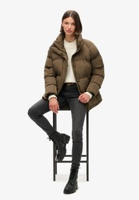 Brun pufferjakke med høj krave, sorte jeans og sorte snørebunds støvler. Jakken har en quiltet tekstur og trykknapper.