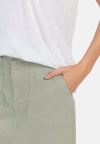 Hellgrau-grüne Hose mit breitem Bund und Seitentaschen, gezeigt mit einer Hand in der Tasche, kombiniert mit einem weißen Oberteil.
