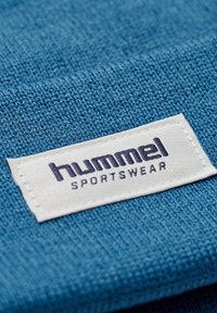 Niebieska dzianina z białą wplecioną metką z napisem "hummel SPORTSWEAR" w ciemnym niebieskim. Gładka tekstura, staranne przeszycia.