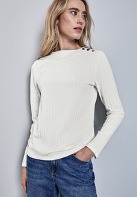 Vit ribbad långärmad topp med en lätt rundad halsringning och tre dekorativa svarta knappar på vänster axel, ihop med blå denimjeans.