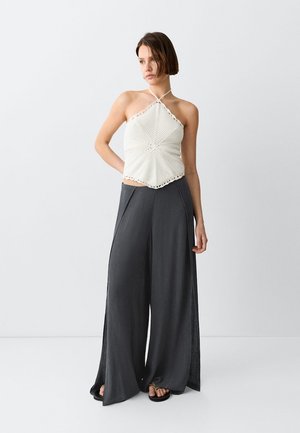 Mujer de pie con un top de crochet blanco tipo halter, pantalones anchos gris oscuro y sandalias negras sobre un fondo liso y claro.
