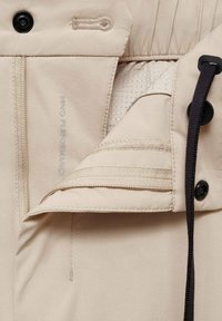 Beige jacket interior with mesh lining, zipper, and button closure. Features elastic bands and branding detail. Smooth texture throughout.