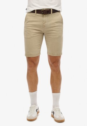Pantalones cortos de algodón beige con un ajuste slim, que cuentan con cremallera y presillas para el cinturón. Un cinturón de piel marrón y zapatillas blancas completan el conjunto.