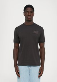 Mann iført en enkel svart t-skjorte med kort erme og et lite rektangulært logo på venstre bryst, kombinert med lyseblå jeans.