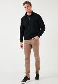 Black zip-up sweatshirt with a high collar, paired with beige slim-fit pants and black sneakers featuring velcro straps.