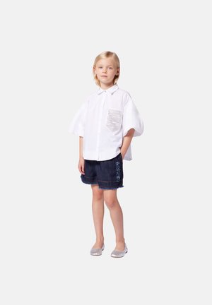 Chemise blanche à manches courtes avec poche et texte, associée à un short en jean foncé avec bord effiloché et broderie bleue. Chaussures argentées.