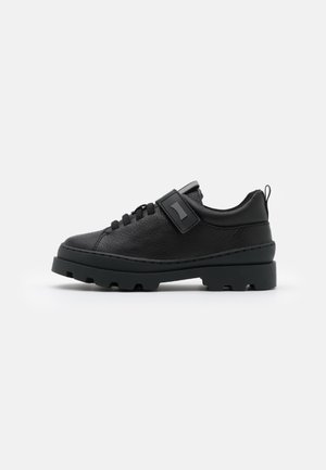 Chaussure en cuir noir avec un bout rond, une semelle épaisse noire et une sangle proéminente sur le dessus. Finition texturée avec des lacets et une languette de tirage.