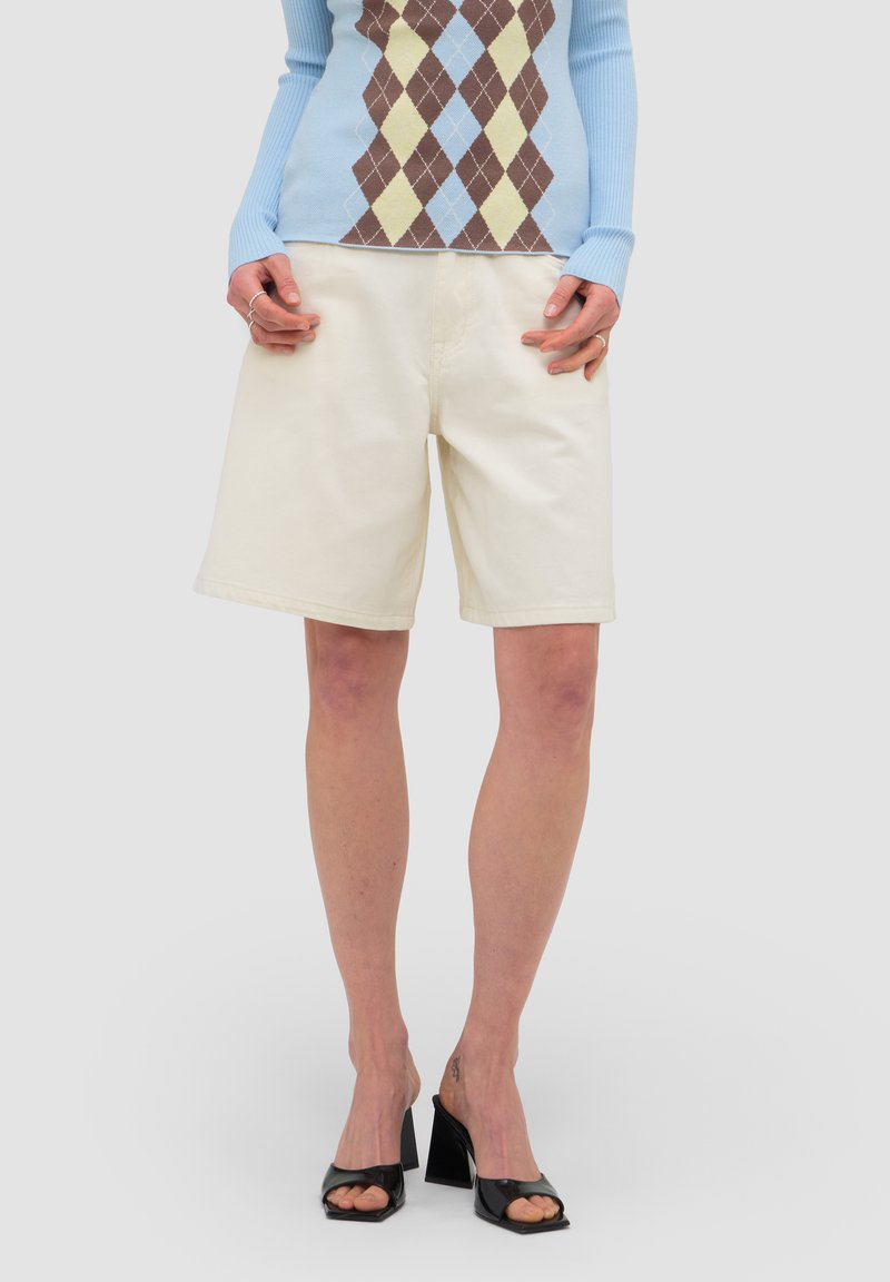 Personne portant un short crème jusqu'aux genoux, des chaussures à talon bloc ouvertes aux orteils noires, et un haut bleu à manches longues avec un motif losanges marron et jaune.