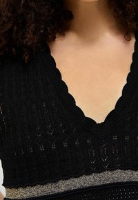 Mujer vistiendo una blusa de punto negra con escote en V y bordes en forma de concha, con una cinturilla metálica dorada, mostrando su cabello rizado y cuello.
