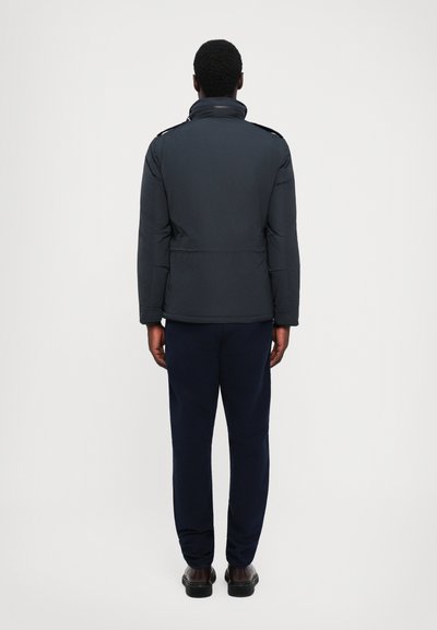 Chaqueta de plumas negra con cuello alto, detalles de cremallera en los lados y un ajuste estructurado, combinada con pantalones azul marino y zapatos oscuros.