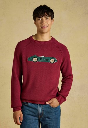 Giovane uomo che indossa un maglione rosso con un disegno di un'auto da corsa vintage verde, in piedi con una mano in tasca davanti a uno sfondo semplice.