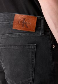 Schwarze Jeans aus Denim mit einem strukturierten braunen Lederpatch, das das Logo "Calvin Klein" zeigt. Verfügt über eine einzelne Gesäßtasche und Gürtelschlaufen.