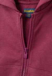 Felpa con zip color marsala, caratterizzata da una zip anteriore, una texture morbida e un'etichetta con la dicitura "essentials" che riporta la taglia e le istruzioni per la cura.