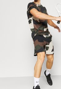 Kombinezon sportowy w kamuflażu, składający się z krótkich rękawów i spodenek w odcieniach zieleni, czerni i beżu; nosiciel w pozie do gry w tenisa.