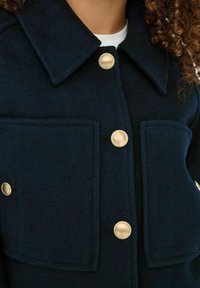 Marine uldjakke med krave, store påsyede lommer og guldtonede knapper. Stoffet har en glat, tekstureret finish.