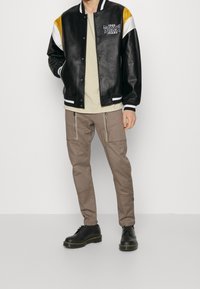 Musta nahkainen varsity-takki, jossa keltaisia ja valkoisia yksityiskohtia, yhdistettynä vaaleanruskeisiin cargo-housuihin ja mustiin nauhakenkiin.