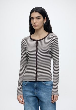 Jeune femme aux longs cheveux foncés portant un haut à manches longues rayé noir et blanc boutonné et un jean bleu, debout devant un fond uni.
