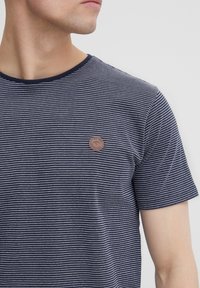 Navy striped t-shirt made of soft fabric with a crew neckline and a small circular logo patch on the left chest, featuring thin white stripes.