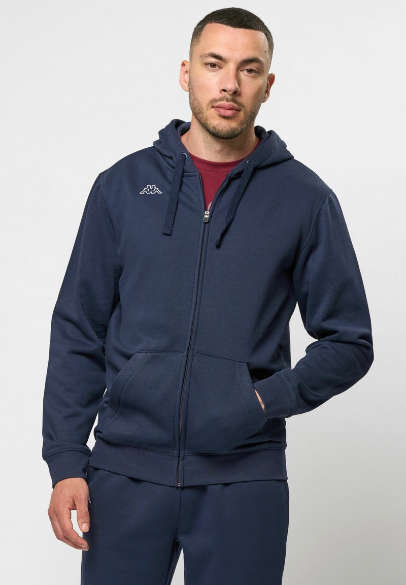 Tumes sinine lukuga kapuutsiga särk, mis on valmistatud pehmest kangast. Omab esiosa taskut, soonikkoes varrukaotsi ja väikest logo rinnal. Ühtlane sile tekstuur.