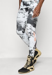 Σφιχτές, γραφικές κολάν σε λευκό και μαύρο με αφηρημένα σχέδια· ελαστικό, διαπνέον ύφασμα. Συνδυασμένες με αθλητικά παπούτσια σε μαύρο και μπεζ.