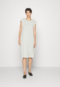 Vestido verde claro, até ao joelho, com mangas curta, feito de tecido suave. Ajuste simples e descontraído, com bolsos laterais. Sapatos planos pretos.