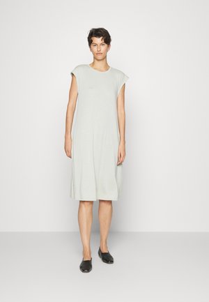 Vestido verde claro, até ao joelho, com mangas curta, feito de tecido suave. Ajuste simples e descontraído, com bolsos laterais. Sapatos planos pretos.