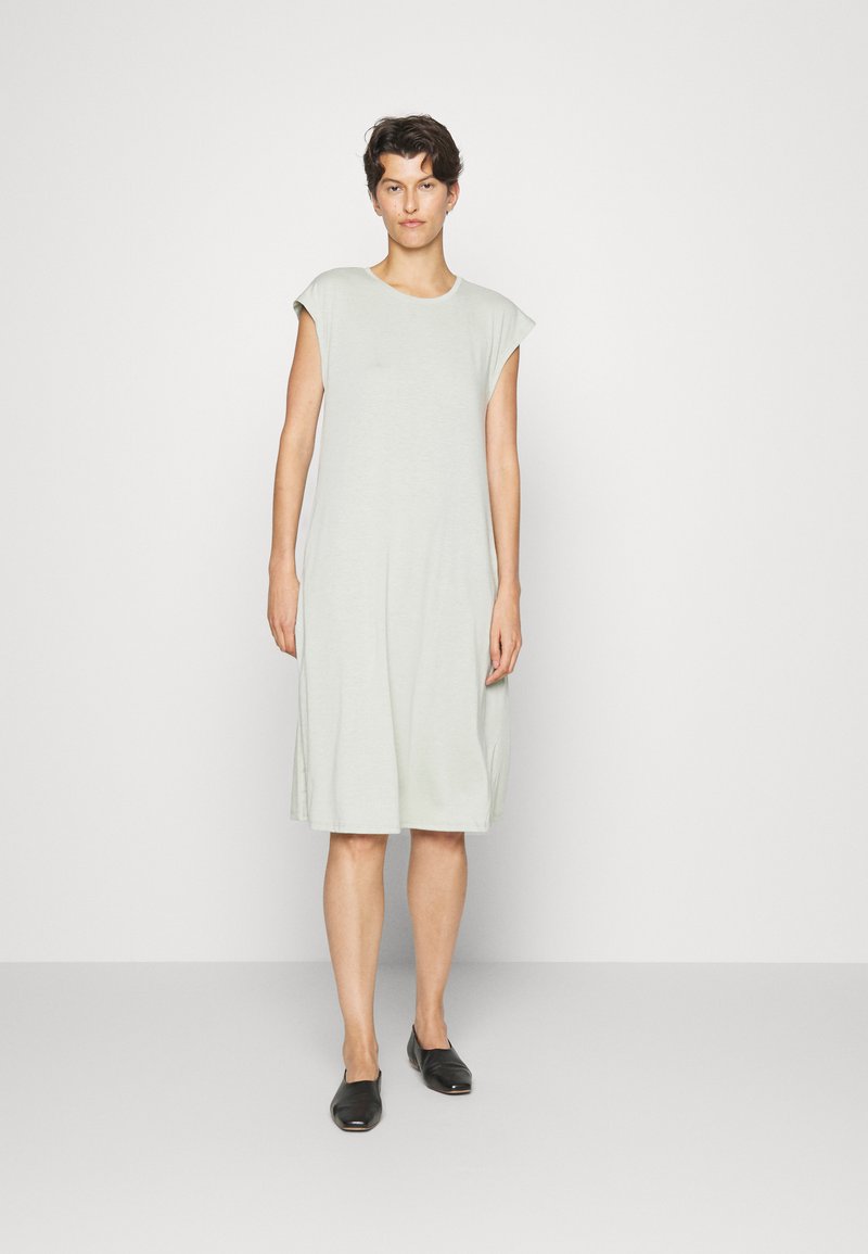 Vestido verde claro, até ao joelho, com mangas curta, feito de tecido suave. Ajuste simples e descontraído, com bolsos laterais. Sapatos planos pretos.