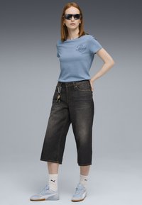Lichtblauw T-shirt met korte mouwen en geborduurd logo, gecombineerd met donkere, cropped denim broek, lichtblauwe sneakers en een zonnebril.