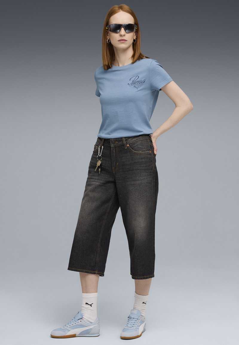 Lichtblauw T-shirt met korte mouwen en geborduurd logo, gecombineerd met donkere, cropped denim broek, lichtblauwe sneakers en een zonnebril.