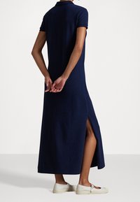 Námořnická modrá bavlněná šaty s krátkými rukávy, polo límcem a bočním rozparkem. Modelka má na sobě světle zbarvené boty. Zobrazeno ze zadní části.