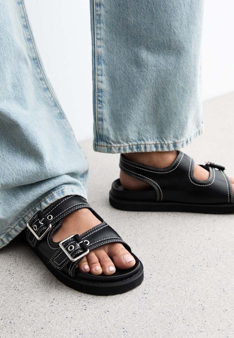 Pieds portant des sandales noires avec des boucles argentées et des coutures blanches, associés à un jean en denim bleu clair sur un sol clair.