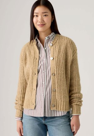 Beige Strickjacke mit strukturiertem Design, rundem Halsausschnitt und fünf Knöpfen, getragen über einem gestreiften Hemd, kombiniert mit hellblauen Jeans.