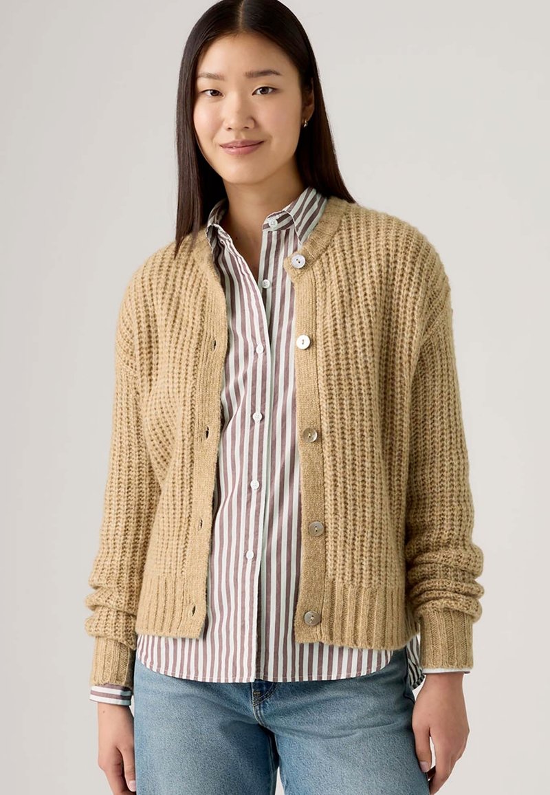 Cardigan lavorato a maglia beige con un design testurizzato, colletto rotondo e cinque bottoni, indossato sopra una camicia a righe, abbinato a jeans azzurri chiari.