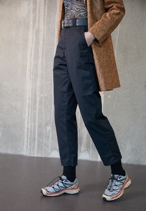 Person trägt schwarze Hose, schwarzes gemustertes Hemd, braune Jacke mit Schlangenprint, schwarzen Gürtel, schwarze Socken und weiße Laufschuhe auf grauem Boden.